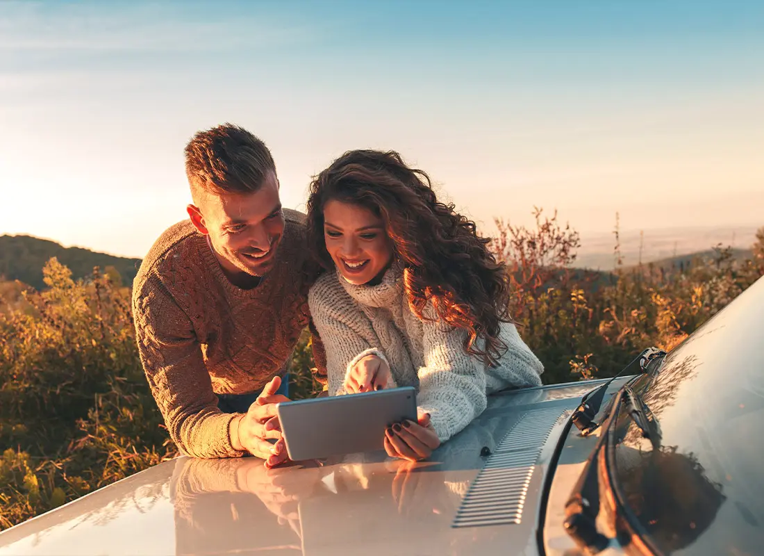 A young man and woman leaning on a car and looking at a tablet together, both smiling for dream assurance service center